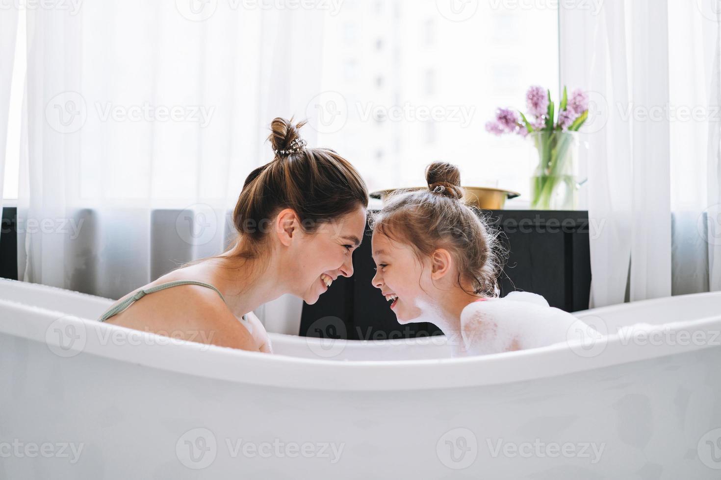 jovem mãe mulher com cabelo comprido com filha adolescente de pijama se divertindo no banho com espuma em casa foto