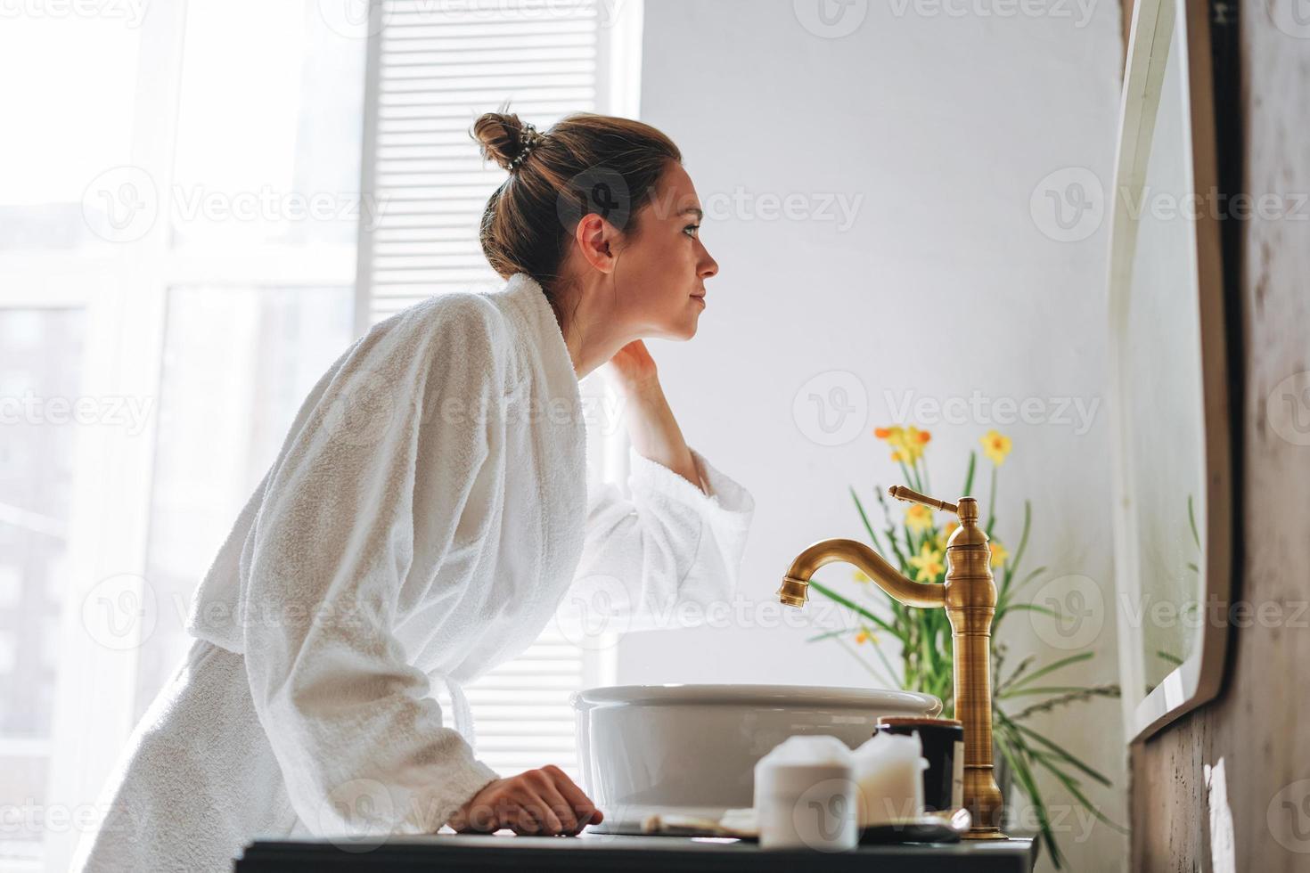 jovem de cabelo comprido escuro em roupão branco perto do espelho no banheiro em casa foto