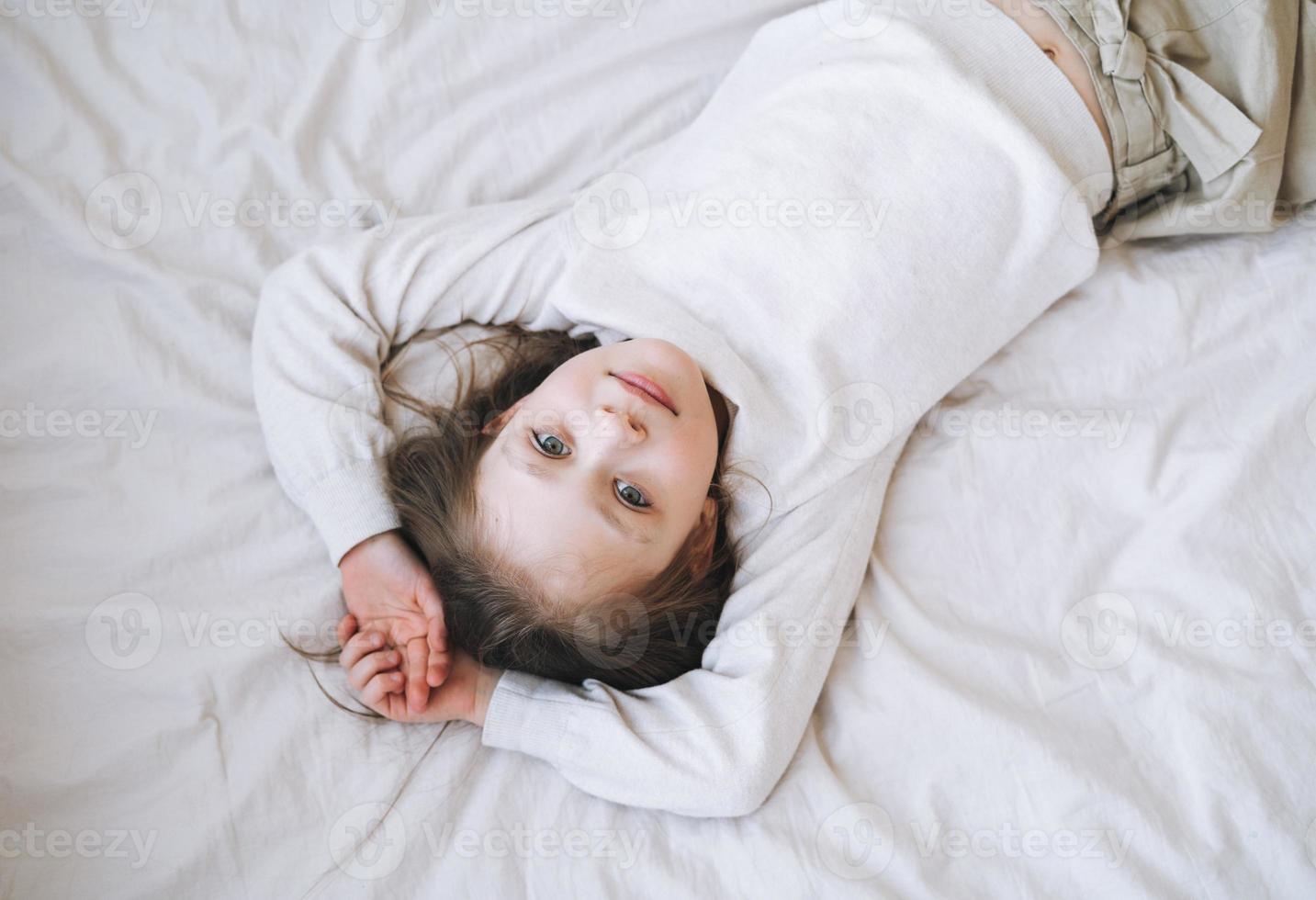 menina engraçada e fofa com cabelo comprido em vestido branco para casa se divertindo deita na cama em casa, vista de cima foto