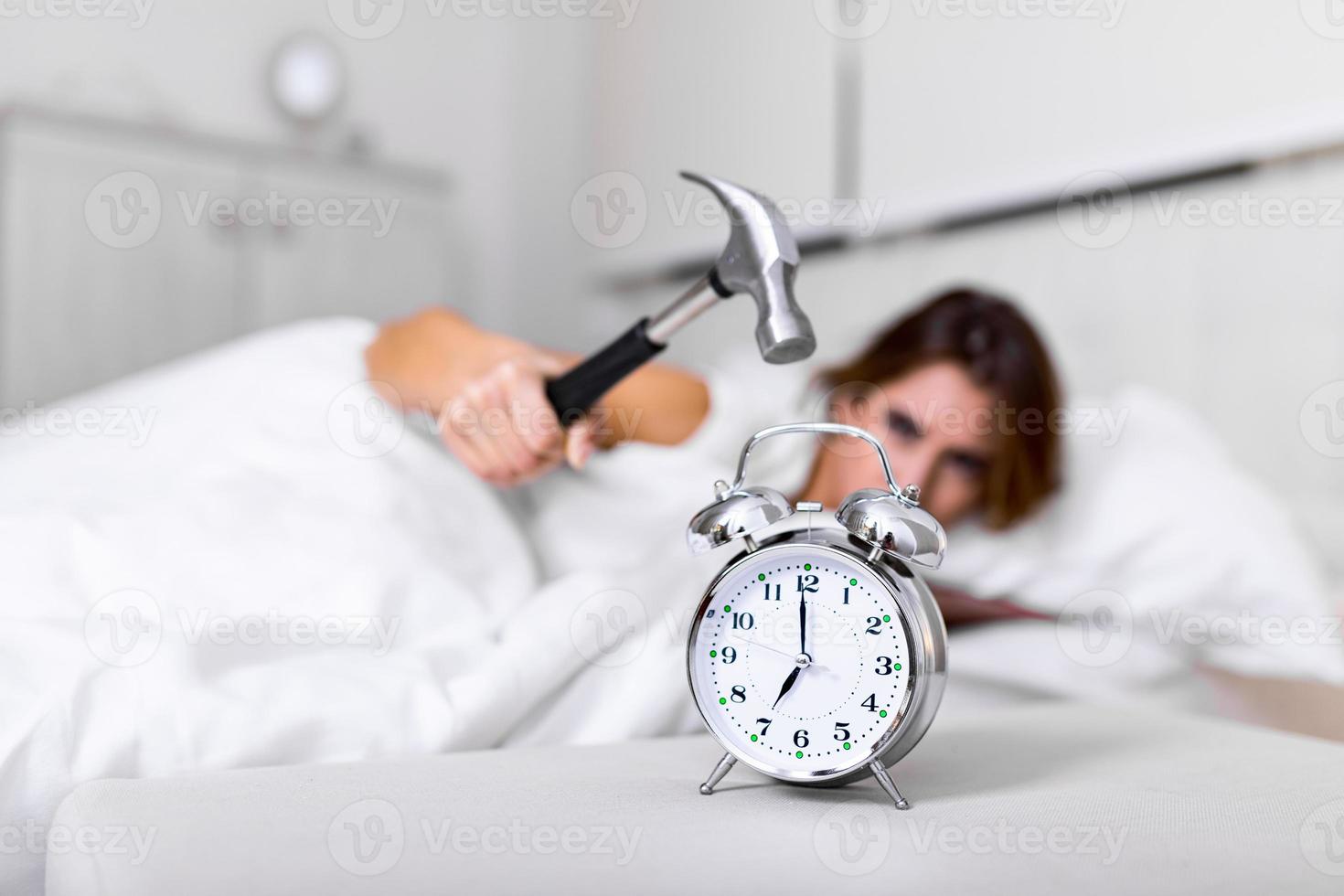 jovem tenta quebrar o despertador com martelo, destruir o relógio. menina deitada na cama desligando um despertador com martelo pela manhã às 7h. foto