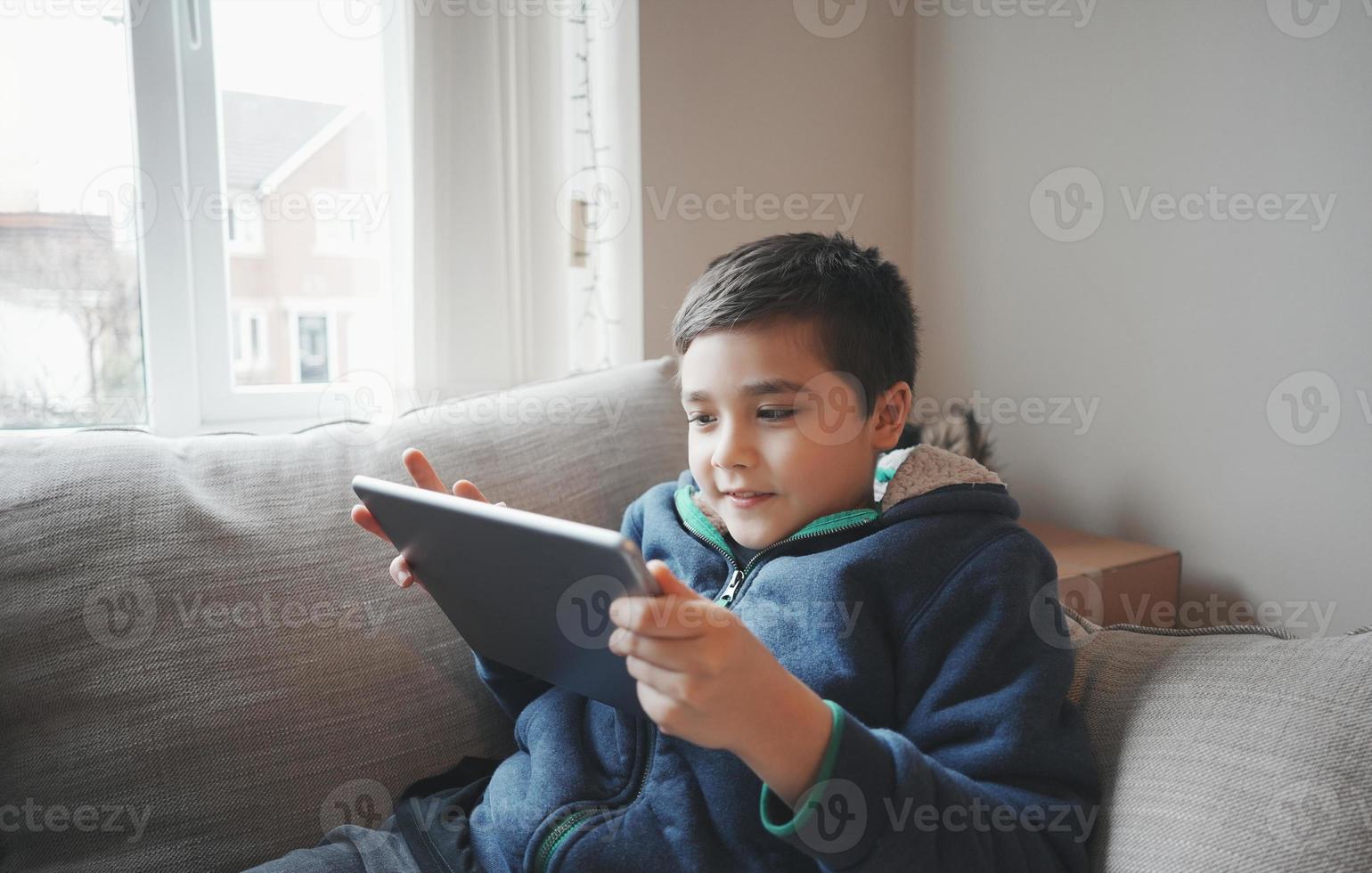 conceito de educação, garoto da escola segurando o tablet lendo e-book para lição de casa, retrato criança feliz jogando jogo online na internet com amigos, menino assistindo desenho animado no teclado digital, garoto sentado no sofá foto