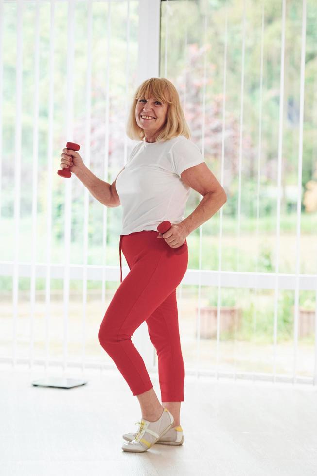 mulher sênior positiva em roupas esportivas dentro de casa durante o dia fazendo exercícios com halteres foto