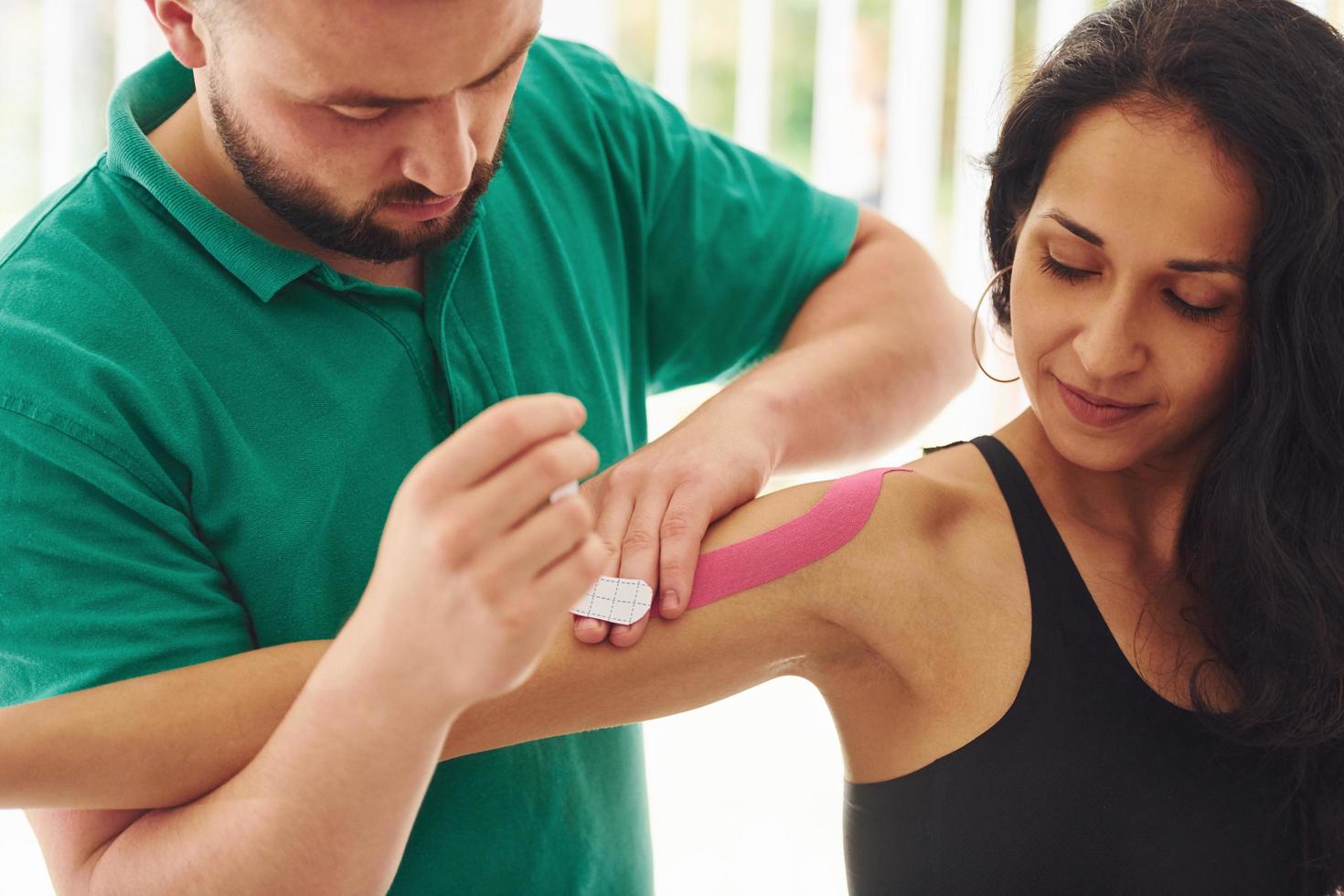 trabalhando com fita fisio. jovem com tipo de corpo fitness tem uma visita na clínica de reabilitação. instrutor ajudando ela foto