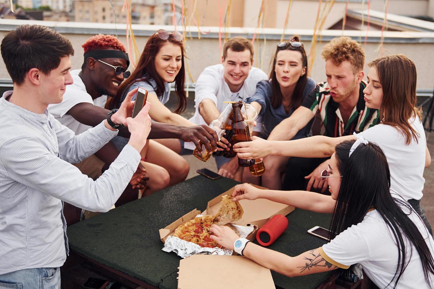 fazendo elogios por garrafas com cerveja. grupo de jovens em roupas casuais faz uma festa no telhado juntos durante o dia foto