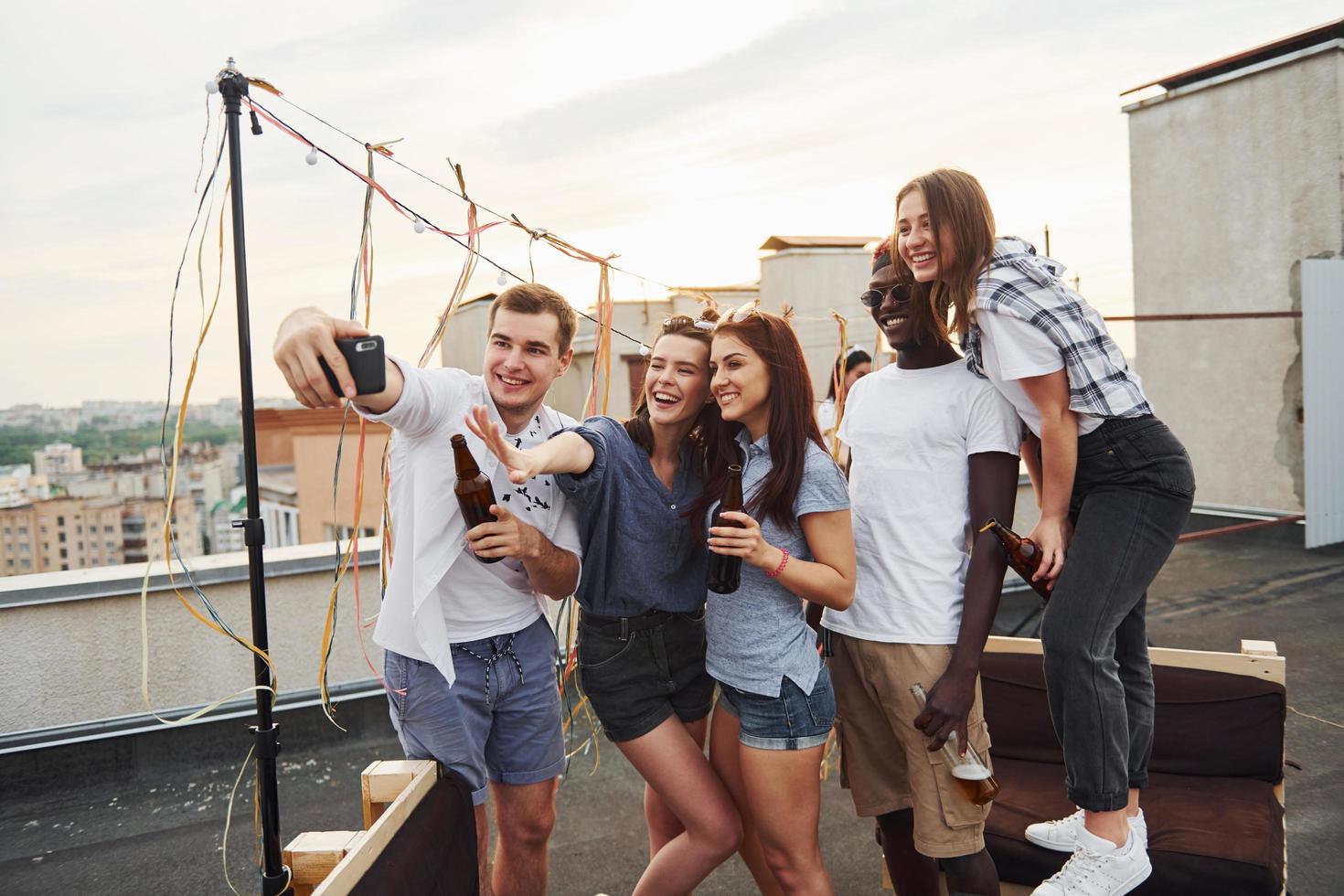 grupo de jovens em roupas casuais faz uma festa no telhado juntos durante o dia e faz selfie por telefone foto