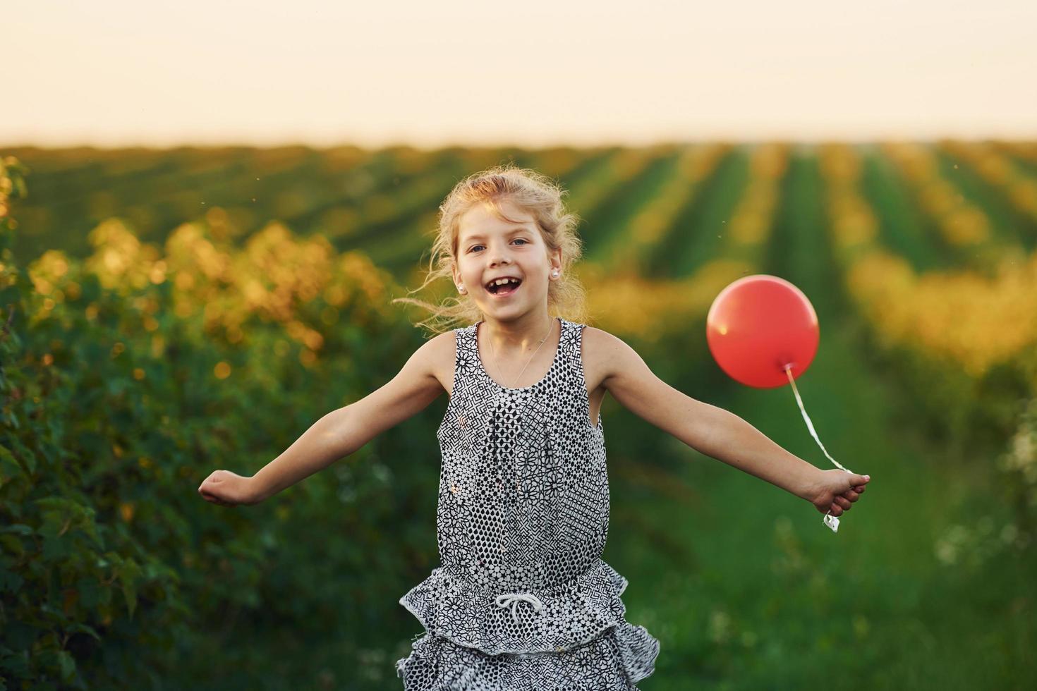 menina positiva com balão vermelho nas mãos divirta-se no campo durante o dia de verão foto