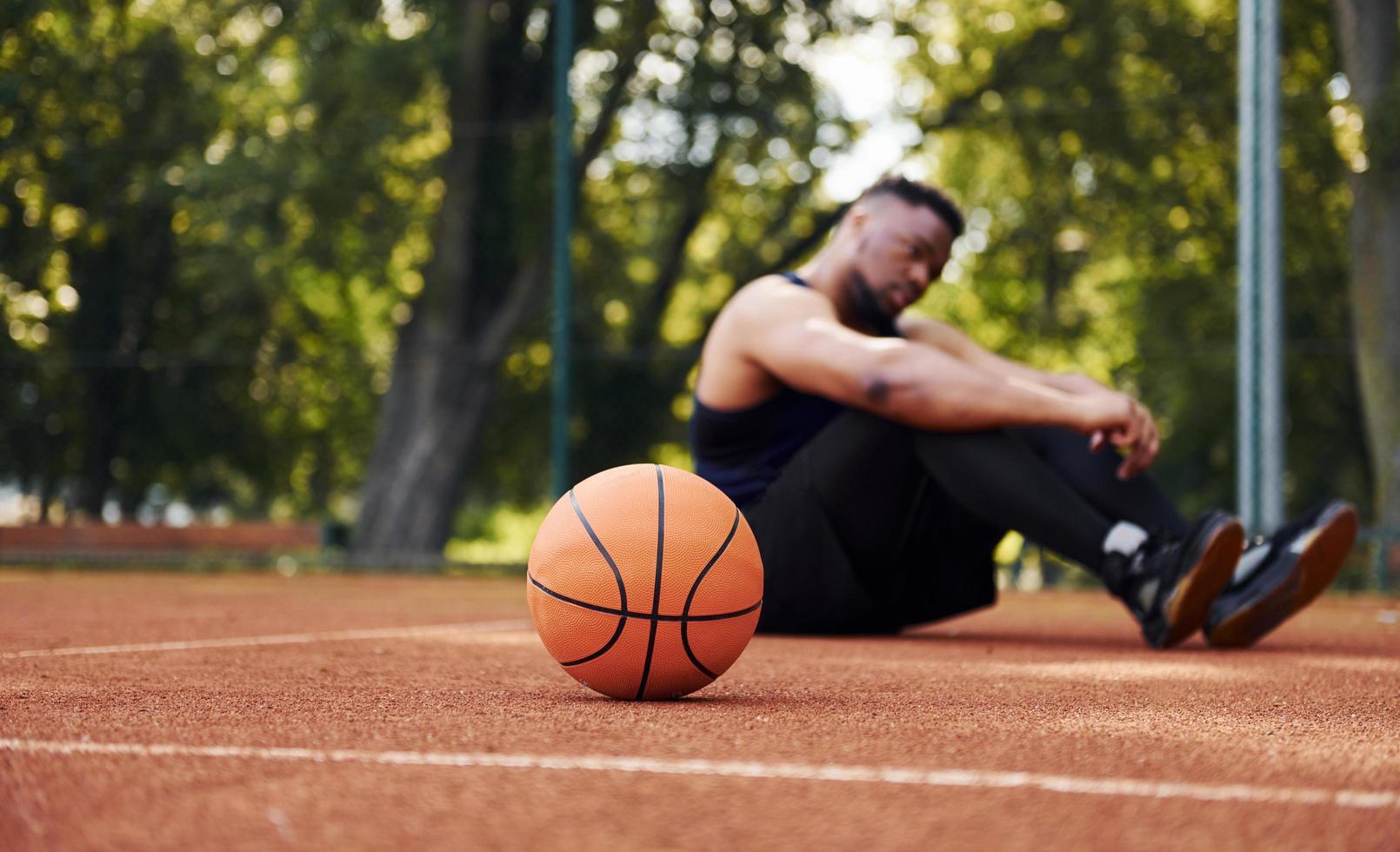 homem afro-americano cansado senta-se no chão com bola na quadra ao ar livre. faz uma pausa foto