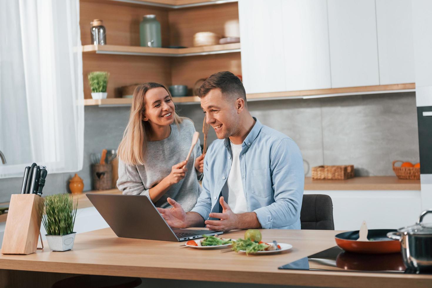 laptop moderno está sobre a mesa. casal preparando comida em casa na cozinha moderna foto