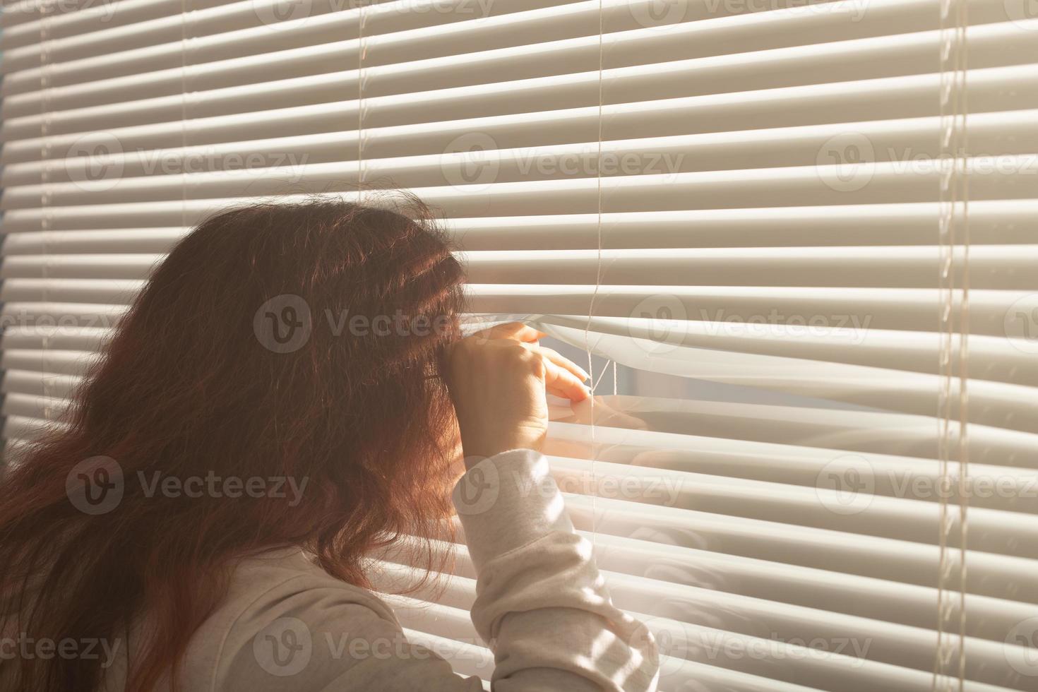 vista traseira da bela jovem com cabelo comprido espreita pelo buraco nas persianas e olha pela janela. conceito de vigilância e curiosidade foto
