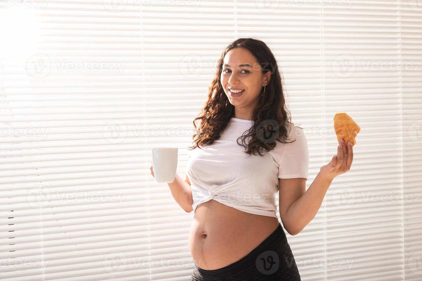 linda mulher grávida segurando croissant e xícara de café nas mãos durante o café da manhã. conceito de boa saúde e atitude positiva enquanto espera o bebê foto