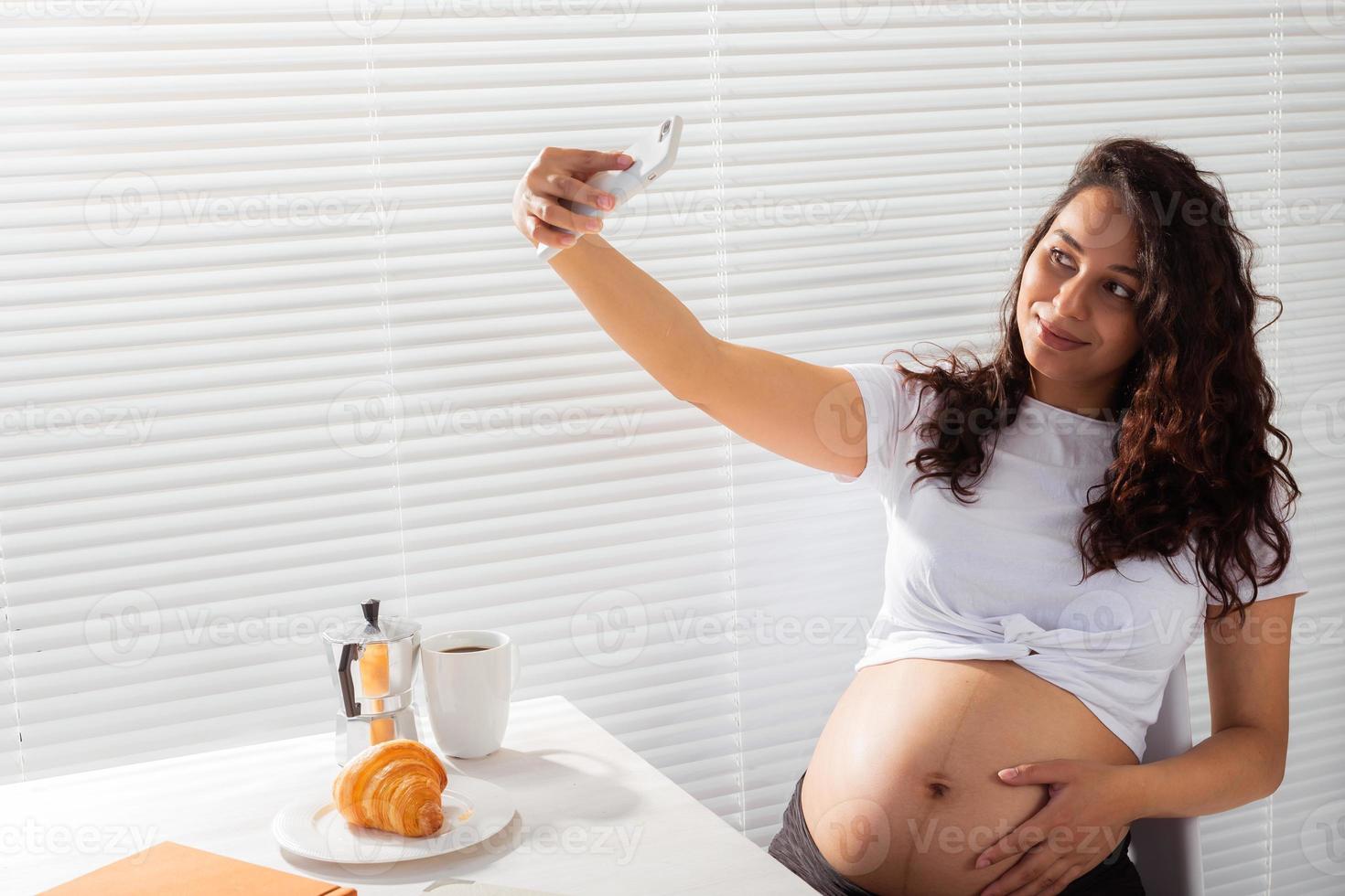 mulher grávida toma selfie em seu smartphone durante o café da manhã. licença de maternidade e conceito de tecnologias. foto