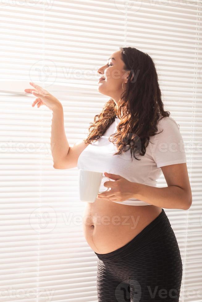 curiosa jovem bonita grávida bebendo chá e olhando através das persianas para a janela. conceito de alegria e boas notícias enquanto espera pelo bebê foto