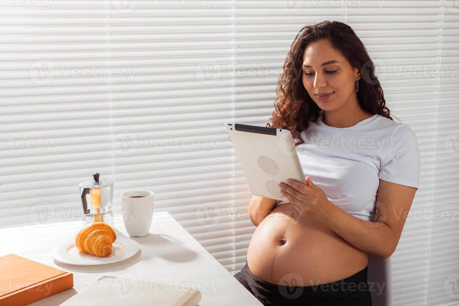 mulher grávida hispânica usando tablet digital durante o café da manhã. tecnologia, gravidez e licença maternidade foto