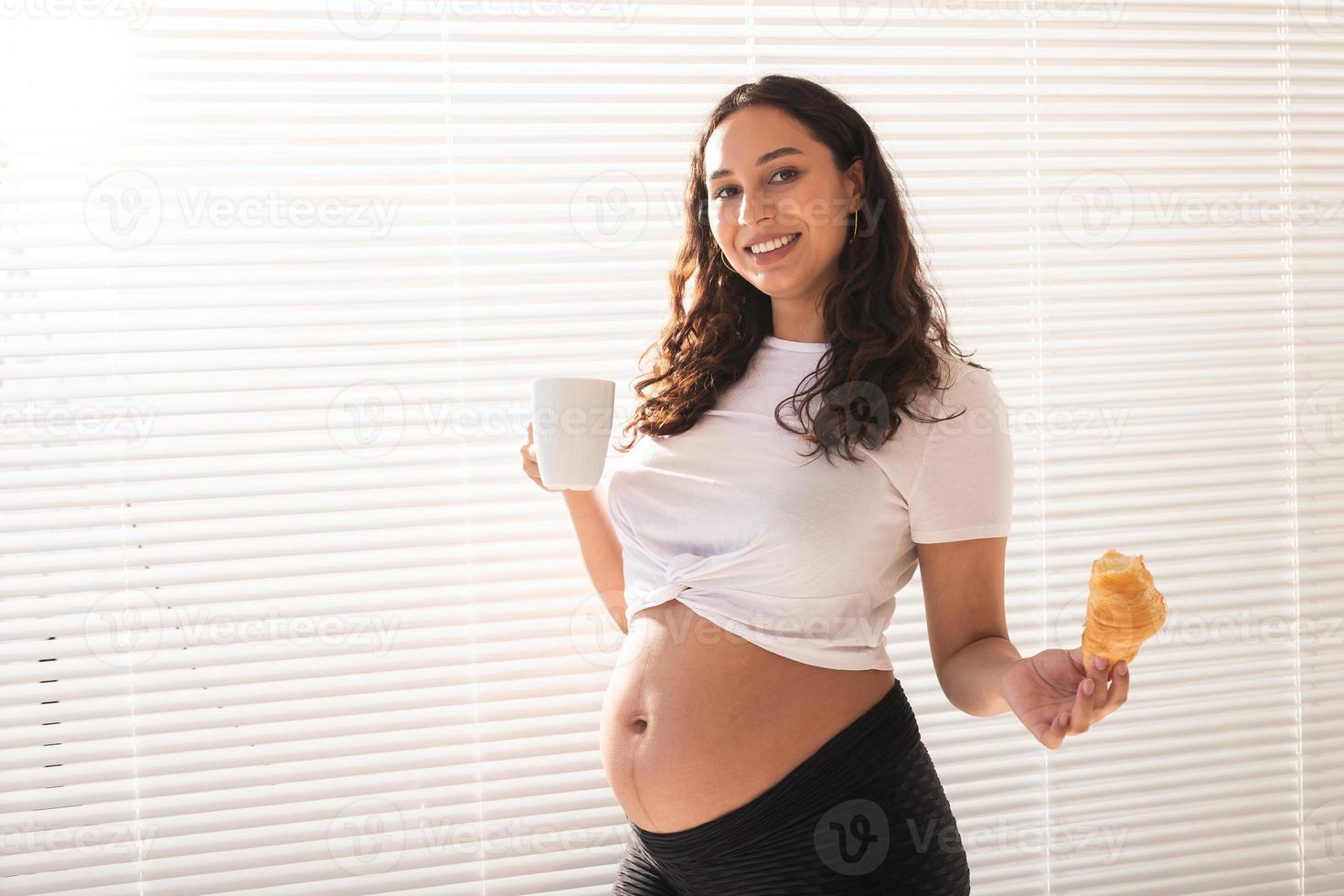 linda mulher grávida segurando croissant e xícara de café nas mãos durante o café da manhã. conceito de boa saúde e atitude positiva enquanto espera o bebê foto