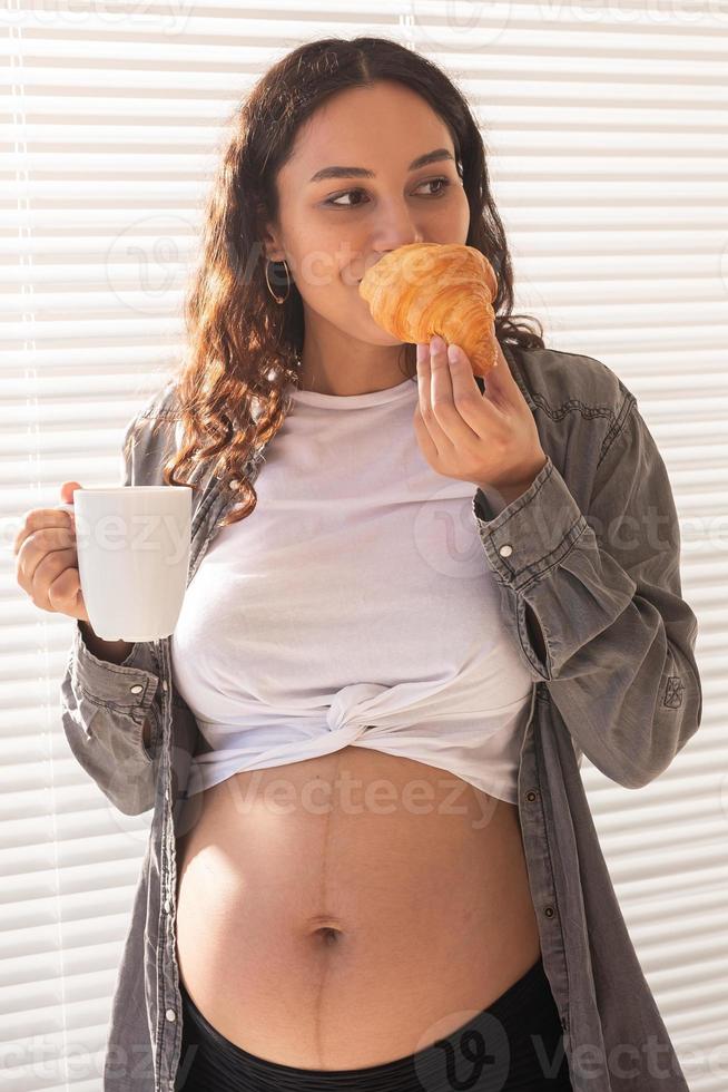 mulher grávida bonita saudável bebendo chá e comendo croissant durante o almoço. conceito de nutrição de alto teor calórico enquanto espera o nascimento do bebê foto