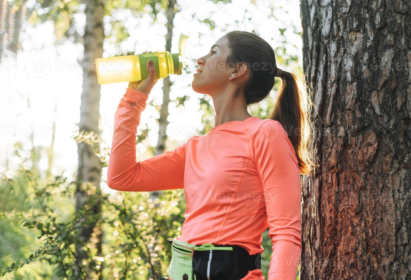 jovem morena magro em roupas esportivas correndo e bebendo água na floresta na hora dourada do nascer do sol. saúde e bem-estar, estilo de vida fitness foto