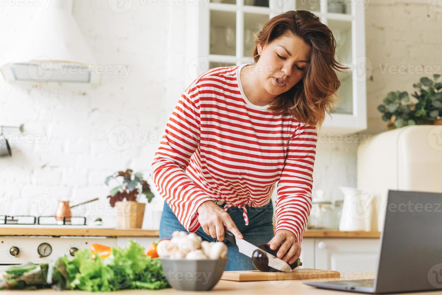 jovem plus size corpo positivo em manga comprida vermelha cozinhando comida usando laptop com receitas de vídeo online na cozinha em casa foto