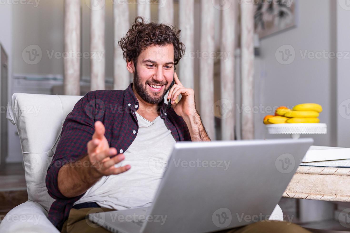 homem bonito sorridente falando no smartphone e usando o computador portátil enquanto está sentado em casa. carreira freelancer. cara falando no celular usando laptop sentado no chão em casa. espaço livre para texto foto