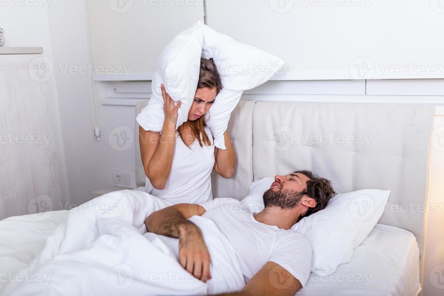 jovem casal tem problema com o ronco do homem. casal heterossexual na cama, homem dormindo e roncando com a boca aberta, enquanto uma mulher cansada irritada por roncar sentada na cama com um travesseiro na cabeça. foto