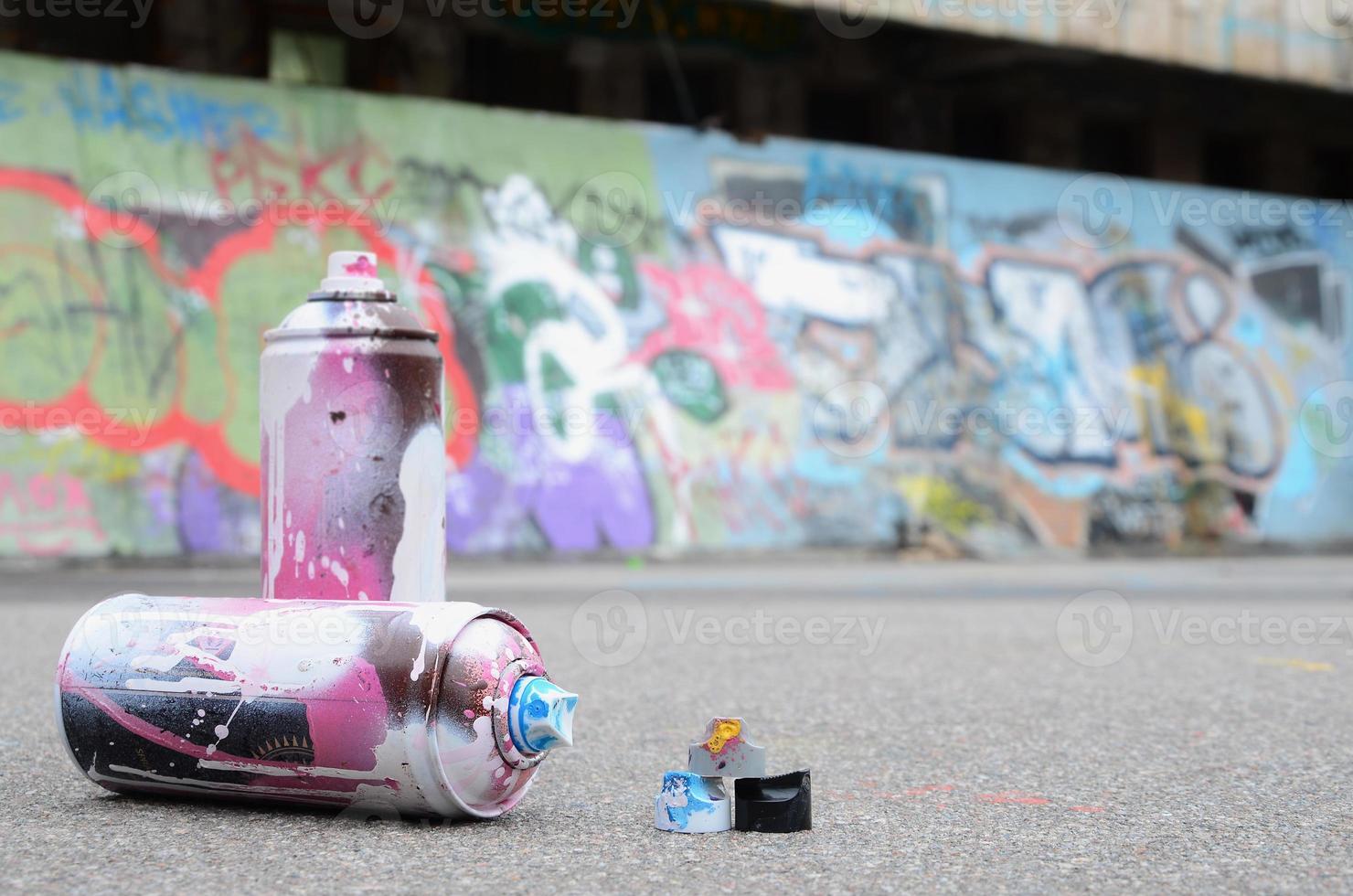 várias latas de spray usadas com tinta rosa e branca e tampas para pulverizar tinta sob pressão estão no asfalto perto da parede pintada em desenhos de graffiti coloridos foto