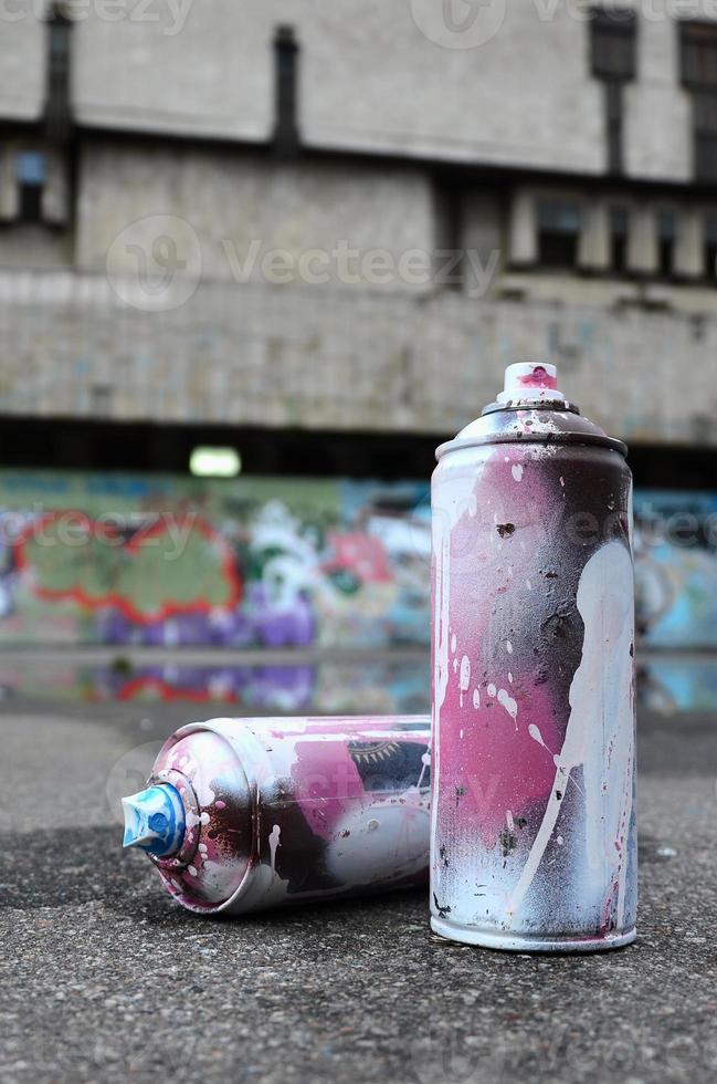 várias latas de spray usadas com tinta rosa e branca e tampas para pulverizar tinta sob pressão estão no asfalto perto da parede pintada em desenhos de graffiti coloridos foto