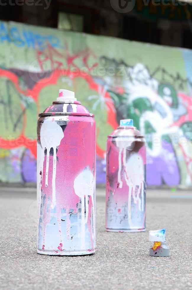 várias latas de spray usadas com tinta rosa e branca e tampas para pulverizar tinta sob pressão estão no asfalto perto da parede pintada em desenhos de graffiti coloridos foto