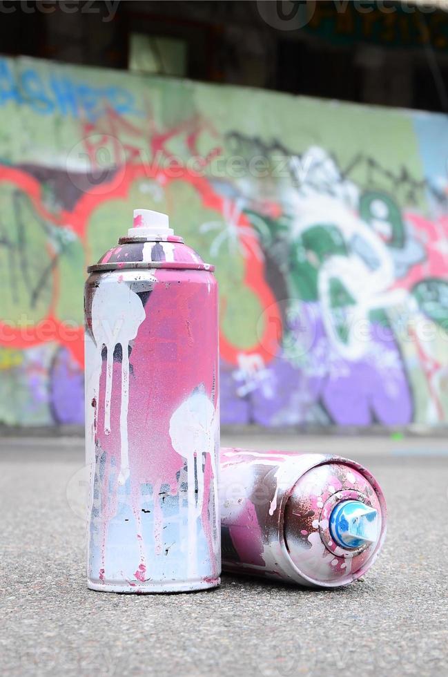 várias latas de spray usadas com tinta rosa e branca e tampas para pulverizar tinta sob pressão estão no asfalto perto da parede pintada em desenhos de graffiti coloridos foto