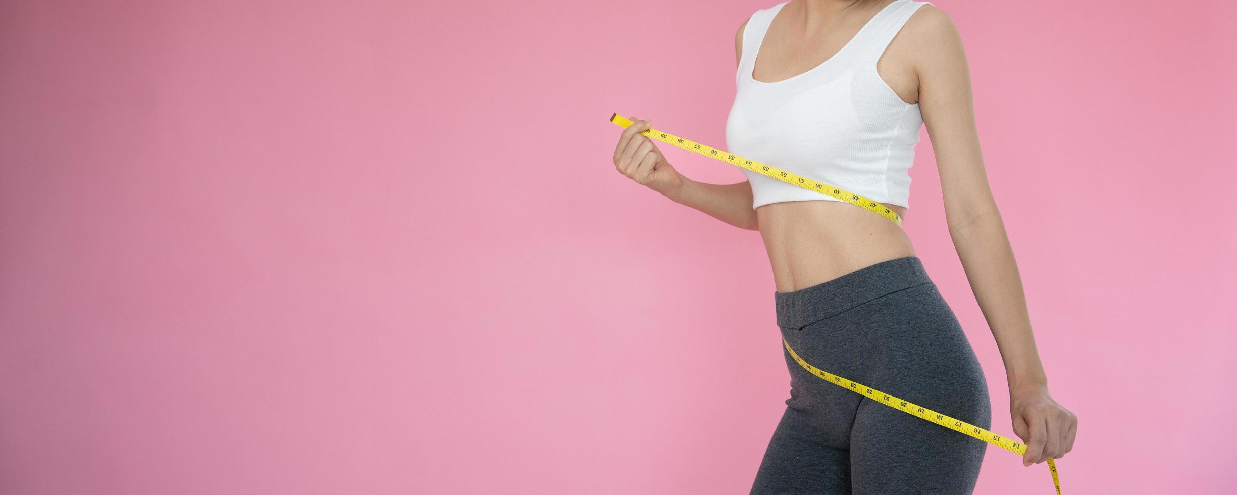 mulher magra em roupas esportivas mede a cintura usando fita métrica no fundo rosa. mulher de dieta e atingir metas de perda de peso foto