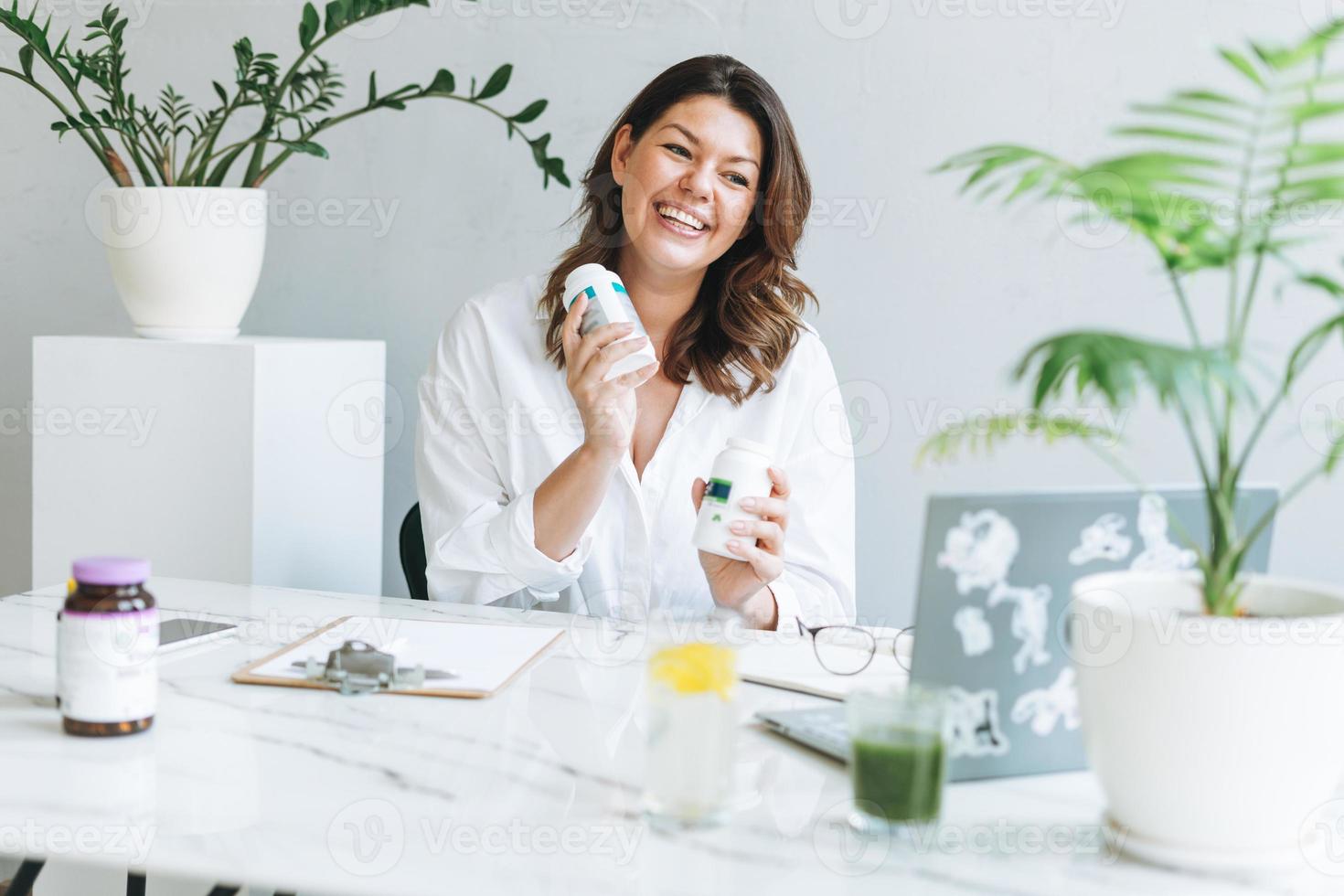 jovem morena sorridente nutricionista plus size em camisa branca trabalhando no laptop na mesa com planta da casa no escritório moderno e brilhante. médico se comunica com o paciente online foto