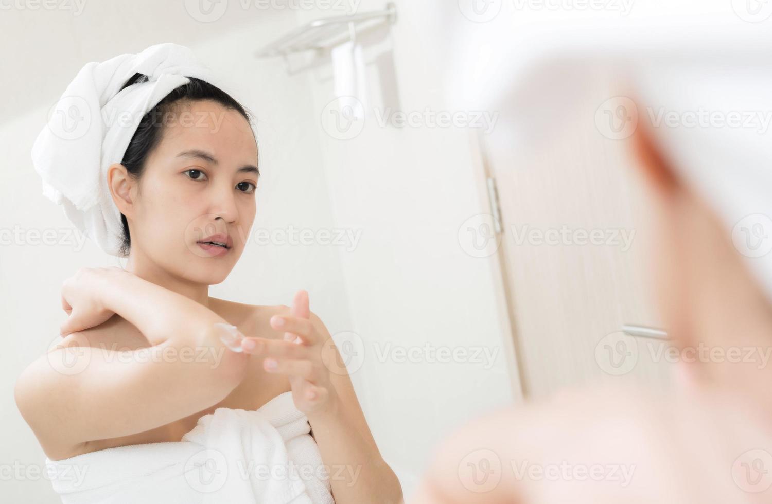 feliz jovem mulher asiática aplicando loções em seu corpo no banheiro foto