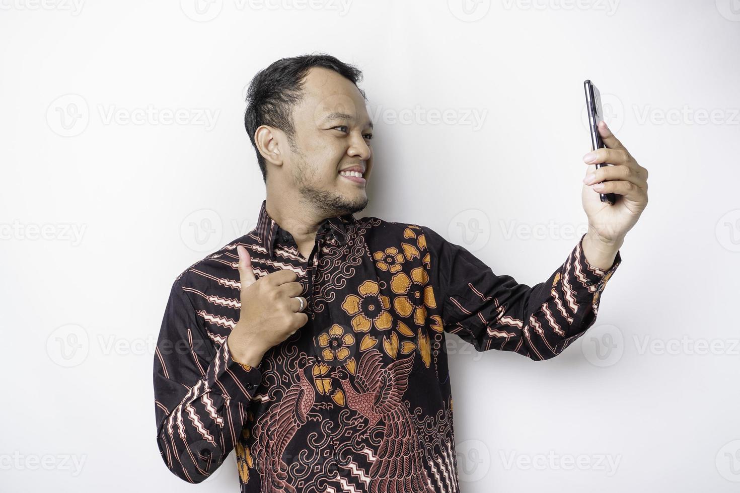 um retrato de um homem asiático feliz vestindo camisa batik e segurando seu telefone, isolado por fundo branco foto