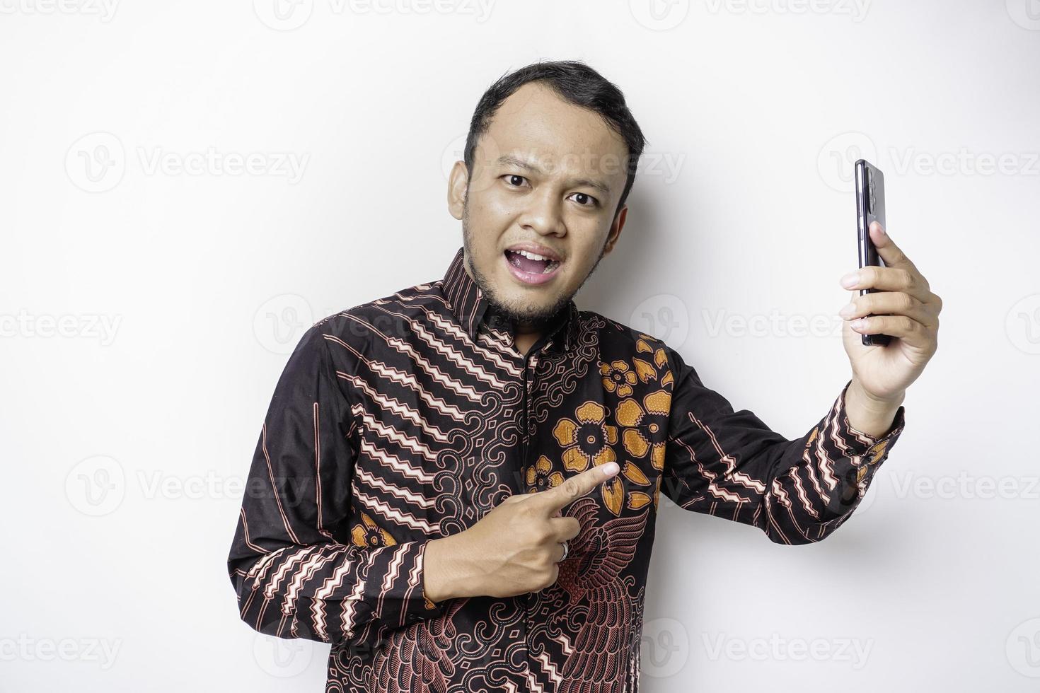 homem asiático chocado vestindo camisa batik e segurando seu telefone, isolado por fundo branco foto