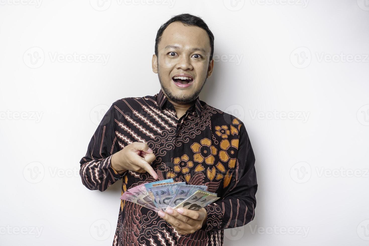 um jovem asiático feliz está vestindo camisa batik e segurando dinheiro em rupias indonésias isolado por fundo branco foto
