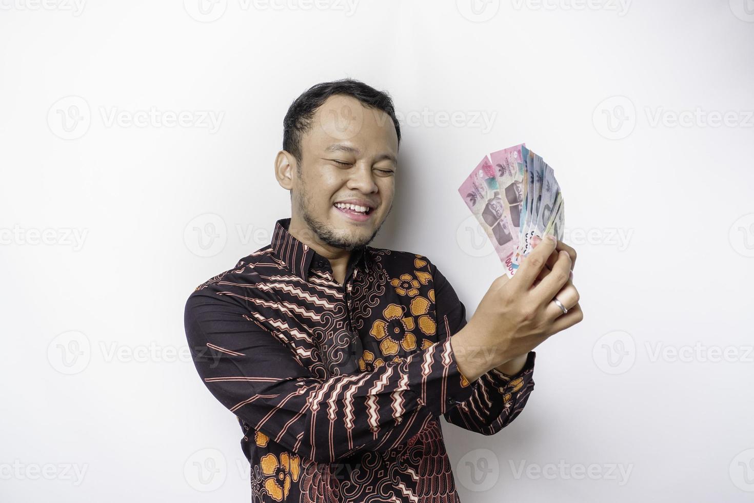 um jovem asiático feliz está vestindo camisa batik e segurando dinheiro em rupias indonésias isolado por fundo branco foto