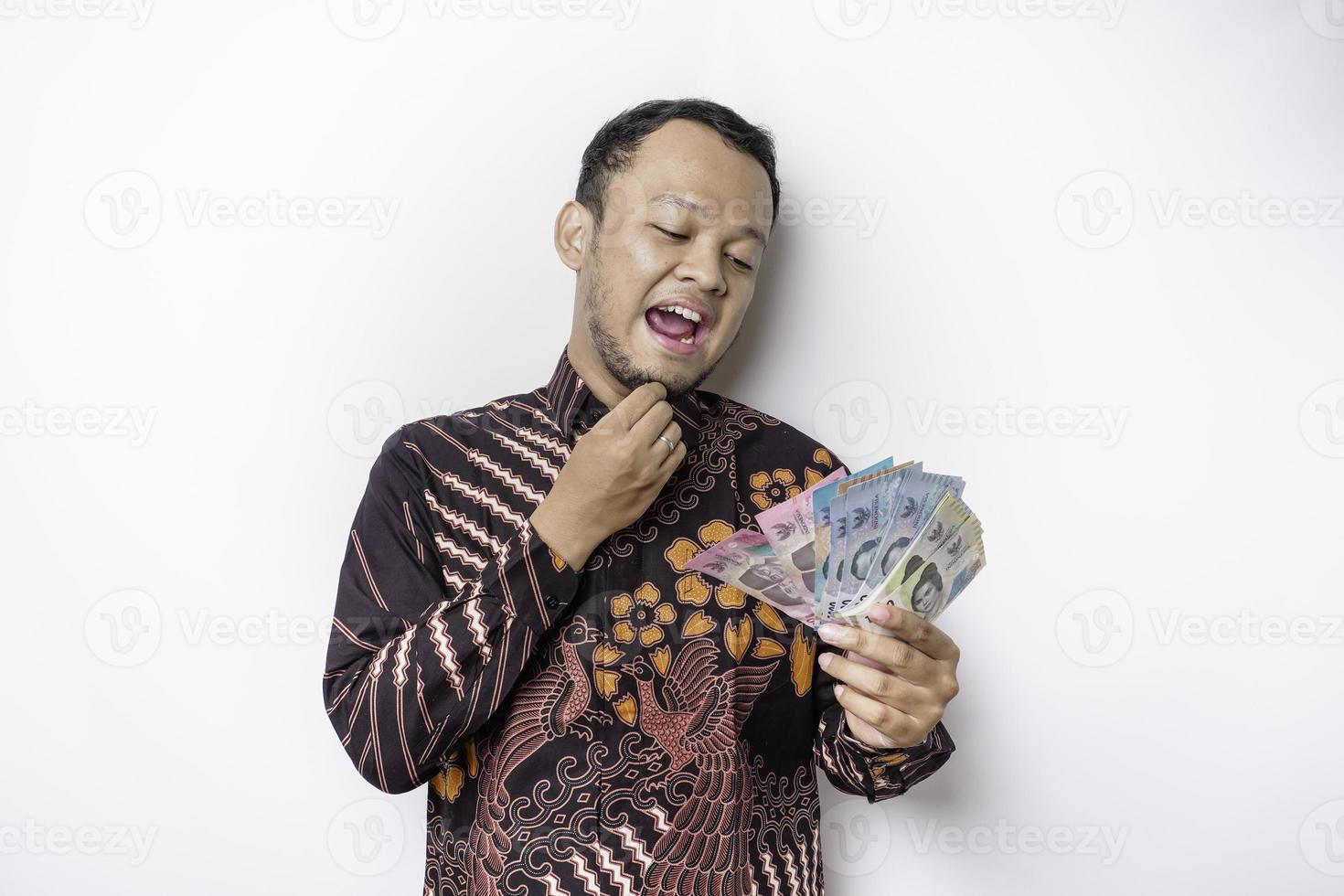 um jovem asiático pensativo está vestindo camisa batik e segurando dinheiro em rupias indonésias isolado por fundo branco foto