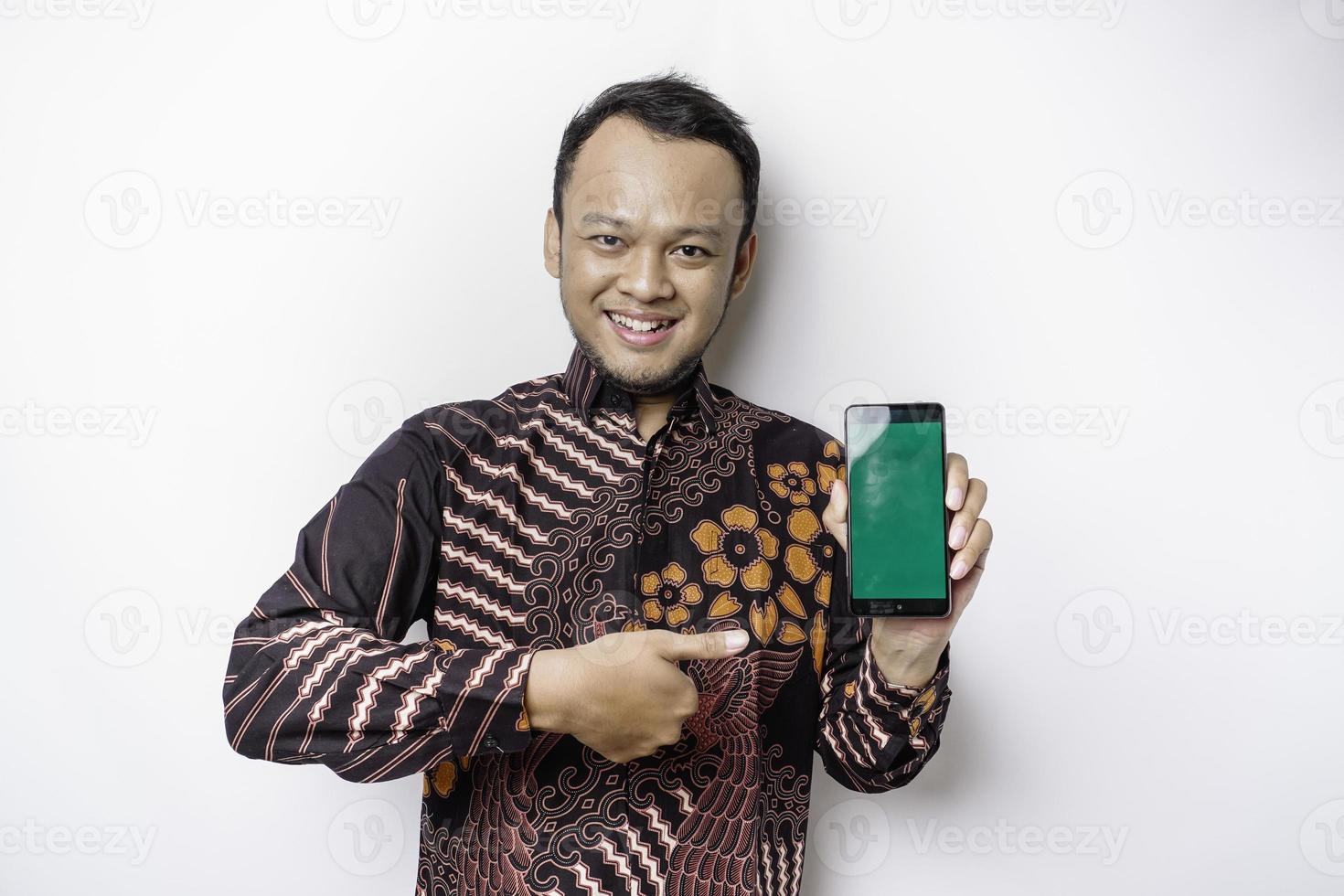 um retrato de um homem asiático sorridente vestindo uma camisa batik e mostrando a tela verde em seu telefone, isolado por fundo branco foto