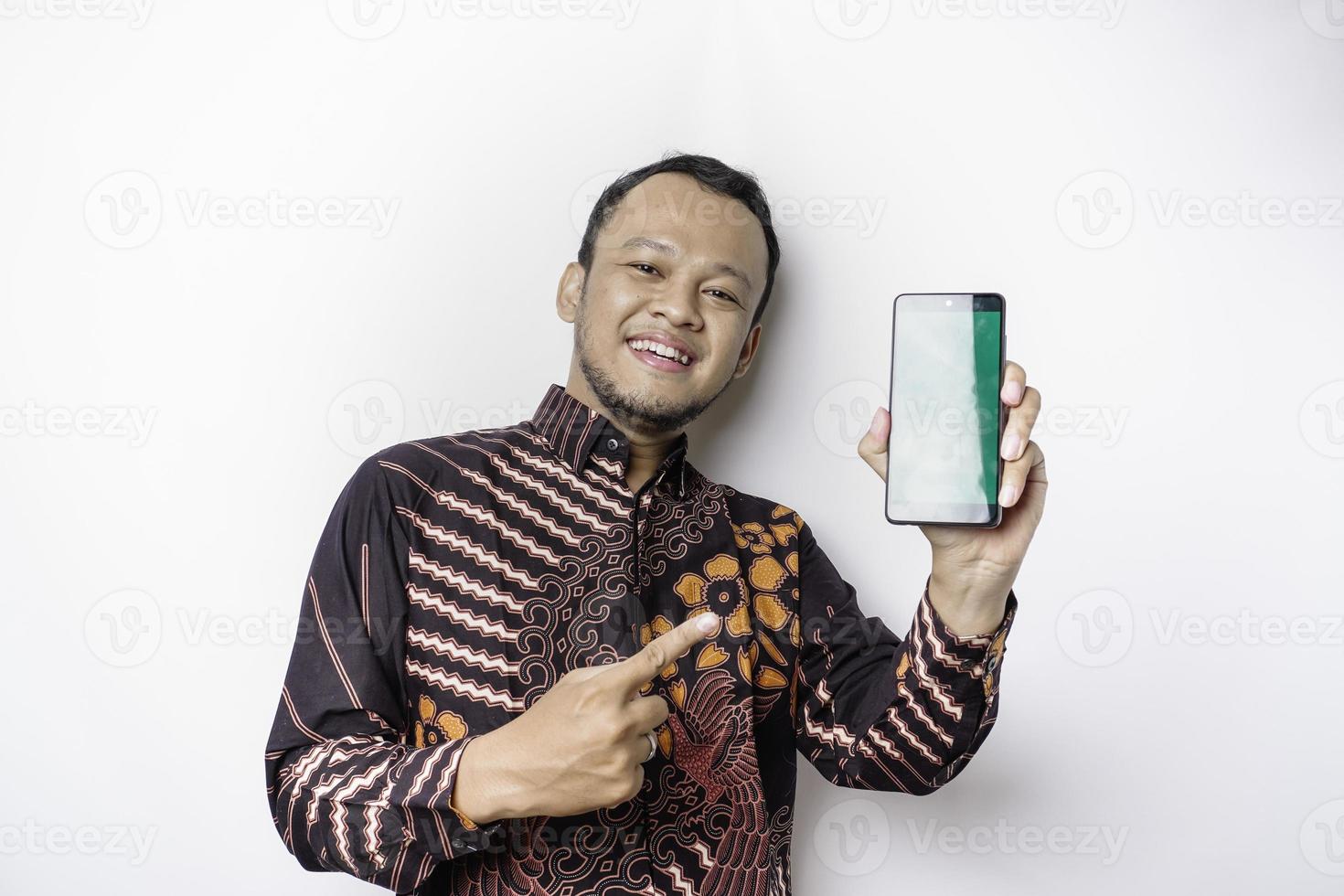 um retrato de um homem asiático sorridente vestindo uma camisa batik e mostrando a tela verde em seu telefone, isolado por fundo branco foto