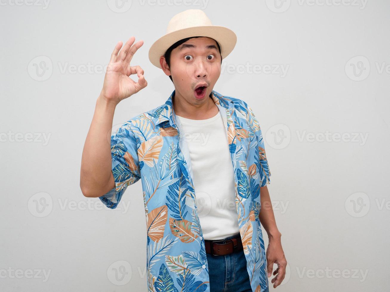 homem asiático de turismo usando camisa de praia de chapéu alegre mostra mão ok isolado foto