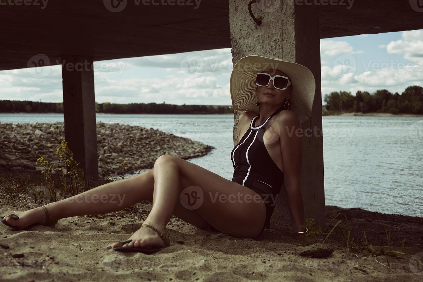 mulher de maiô, chapéu e óculos de sol senta-se na areia no verão na margem do rio em um píer de concreto foto