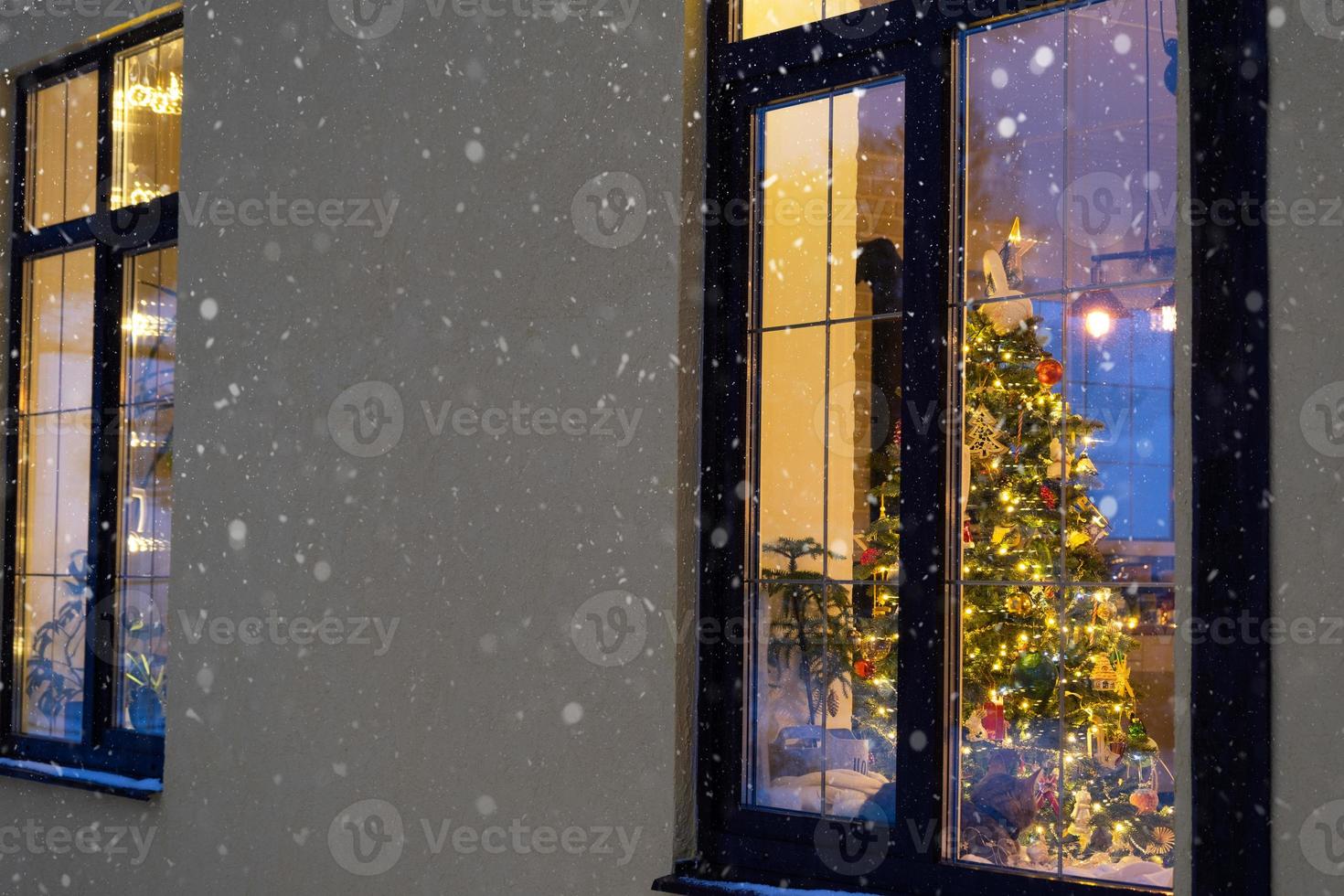 janela festiva aconchegante da casa do lado de fora com a luz quente das guirlandas de luzes de fada dentro - comemore o natal e o ano novo em um lar aconchegante. árvore de natal, bokeh, neve em pinheiros e queda de neve foto