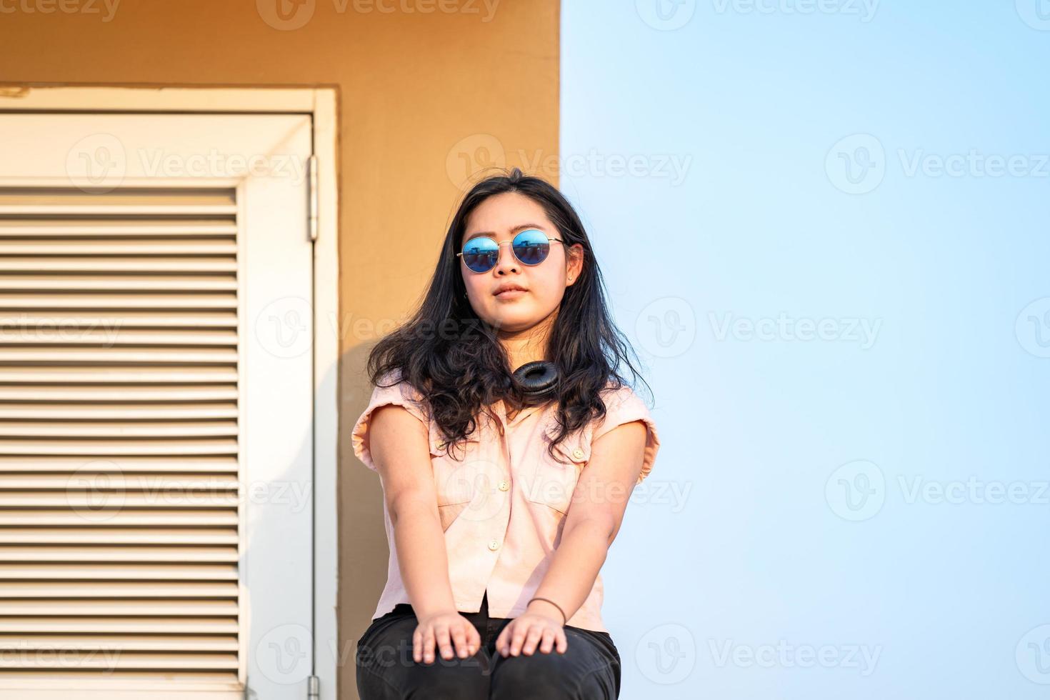 óculos de sol mulher asiática está sentada no cimento barricada do telhado não é perigoso porque o local oposto é o piso do telhado também na frente do céu vazio. foto
