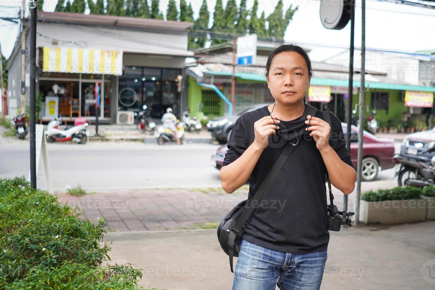 homem asiático de camiseta preta e calça jeans está usando máscara facial preta em frente ao prédio comercial da tailândia e área de estrada rural. foto