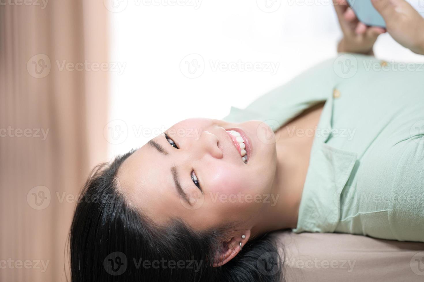 linda mulher asiática está deitada e jogando móvel na cama em seu quarto com um verde pastel - tema de cor marrom. foto