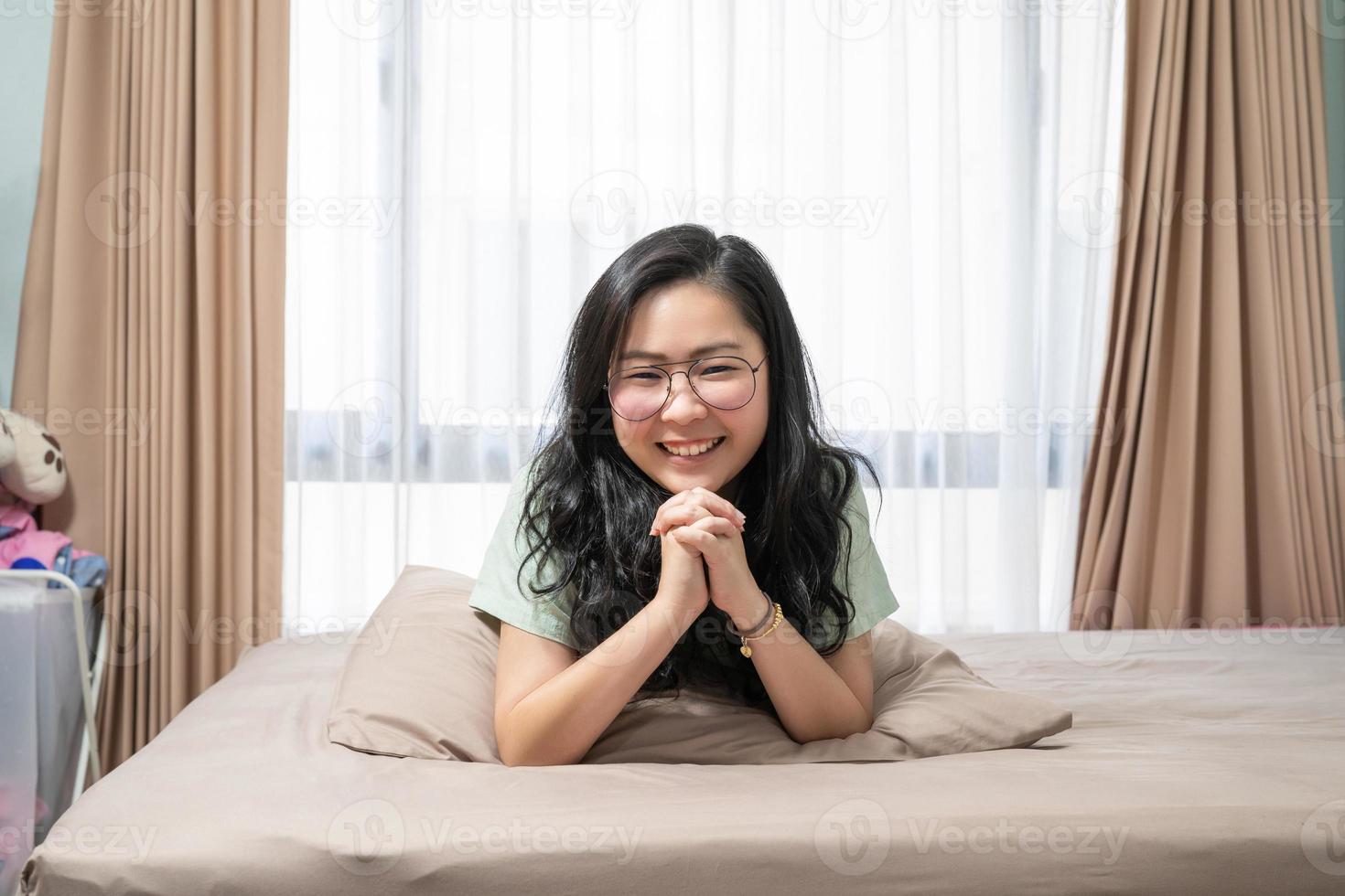 linda mulher asiática mantém as mãos juntas enquanto deita de bruços na cama no quarto com um tema de cor verde pastel - marrom. foto