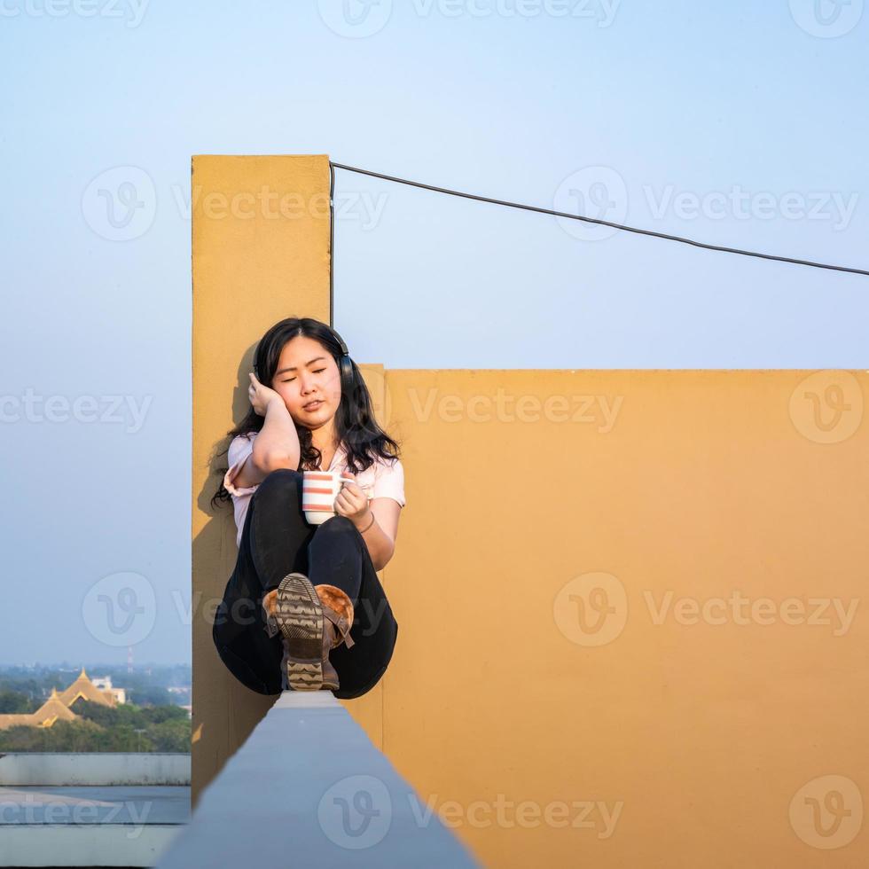 óculos mulher asiática está ouvindo música via fone de ouvido, tomando café no telhado barricada de cimento não é perigoso porque o local oposto também é o último andar. foto