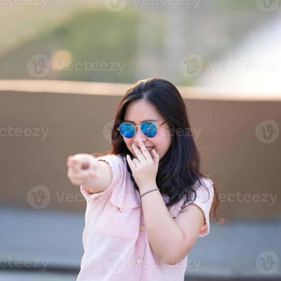 mulher asiática de camisa rosa está jogando arma de dedo, mire e concentre-se nela. ela relaxa e sorri entre as atuações. o ambiente ao redor da construção do telhado no crepúsculo. foto