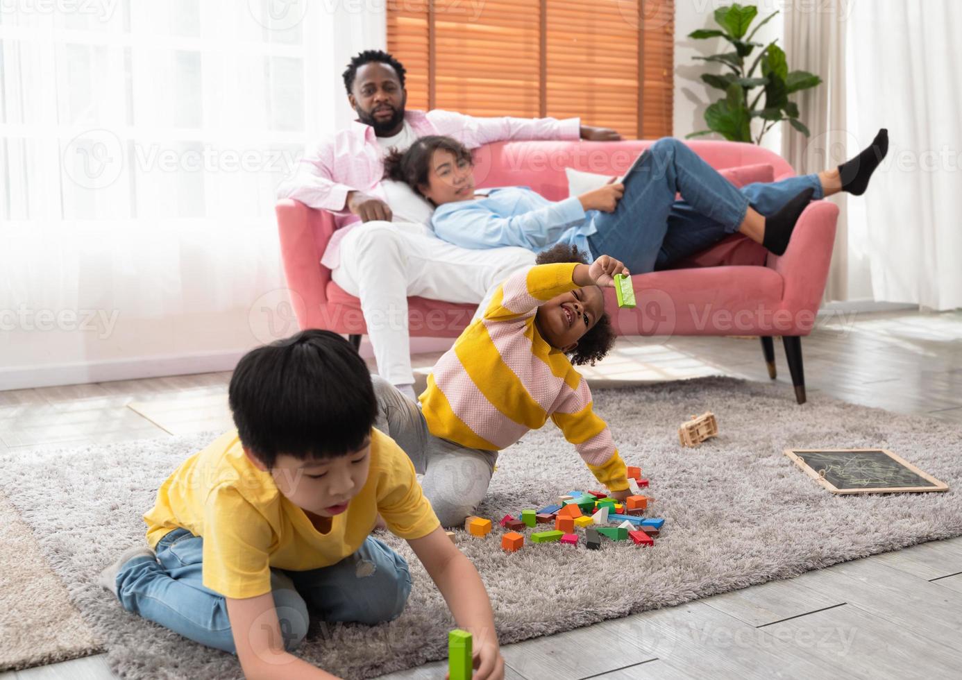 crianças felizes brincando com brinquedos e curtindo a atividade de recreio enquanto estão sentados no tapete do chão na sala de estar. casal negro de pais descansando no sofá e observando as crianças foto