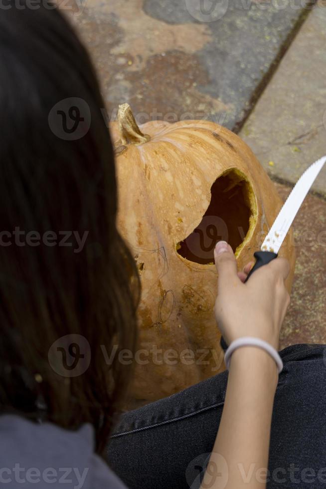 fêmea esculpindo grande abóbora laranja para o halloween enquanto está sentado na mesa de madeira em casa foto