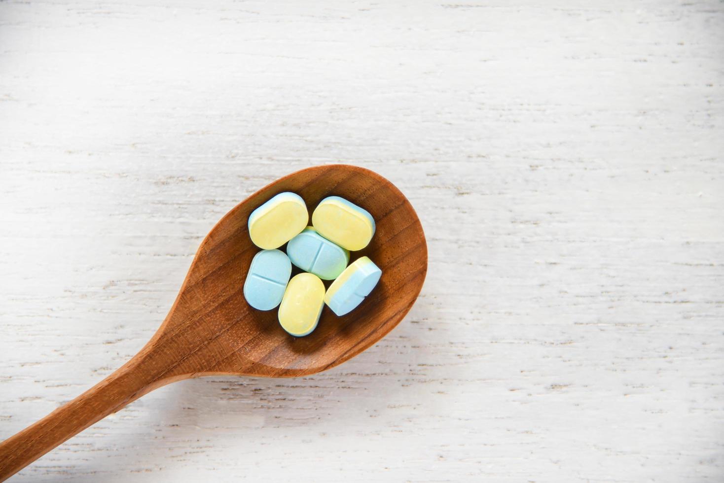 pílulas de remédio amarelas e azuis em colher de pau no fundo da mesa de madeira branca em farmácia foto