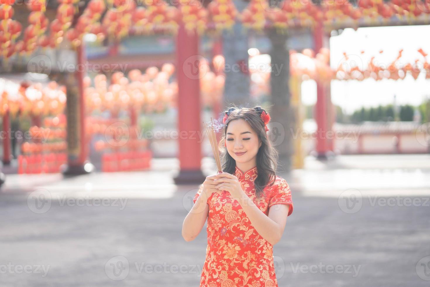 mulher asiática usando vestido cheongsam qipao tradicional rezando com incensos para bênçãos de desejo e boa sorte no templo budista chinês. sorriso de emoção foto